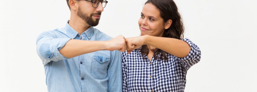 Satisfied couple making fist bump gesture. Young woman in casual and man in glasses standing isolated over white background. Cooperation concept
