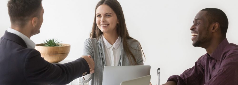 Smiling millennial businesswoman shaking hand of male partner at diverse group negotiation, friendly woman boss handshaking new team member at meeting, showing respect or making contract deal concept